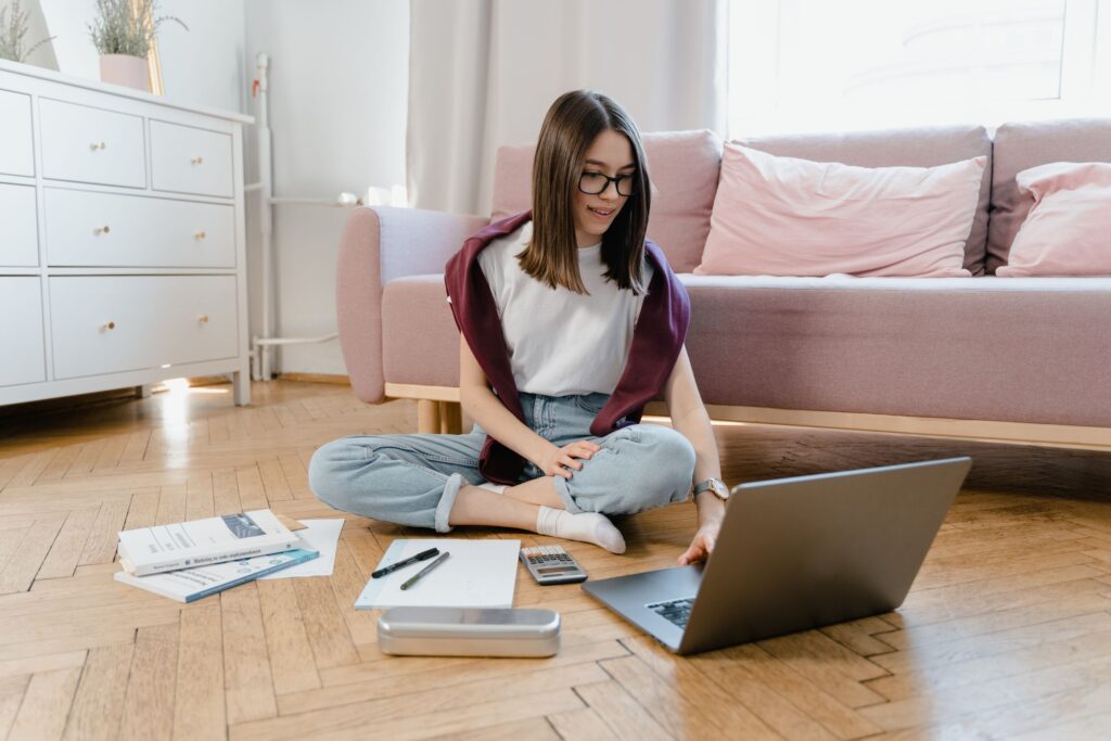 Teenage student sitting on the living room floor working on a laptop with notes and books around her